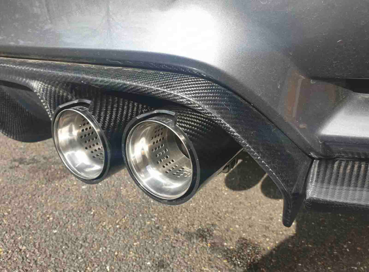 Car exhaust pipes made of shiny metal protrude from a carbon fiber rear diffuser, positioned above a gravel surface, with sunlight reflecting off the vehicle's body.