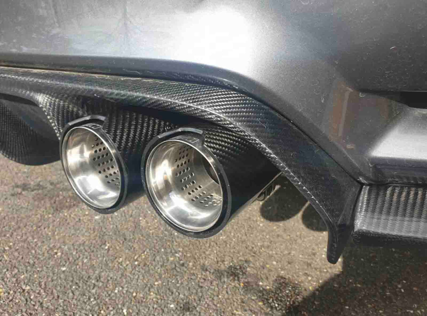 Car exhaust pipes made of shiny metal protrude from a carbon fiber rear diffuser, positioned above a gravel surface, with sunlight reflecting off the vehicle's body.