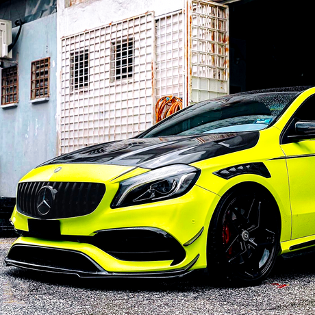 A bright yellow sports car with a glossy black hood is parked in front of a weathered building with barred windows and coiled orange hoses.