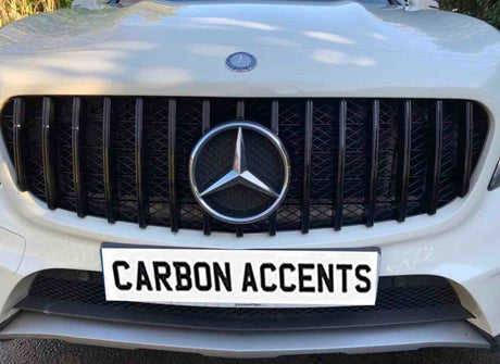 A car grille displays a prominent emblem and black vertical slats, situated on a white vehicle. The license plate reads "CARBON ACCENTS," with greenery faintly visible in the background.