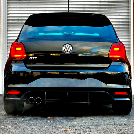 A black Volkswagen GTI is parked, displaying its rear with dual exhausts. Red lights are illuminated, positioned in front of a metallic roller door on a paved surface.
