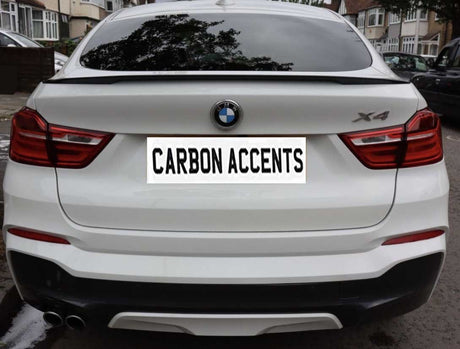 A white BMW X4 is parked on a residential street. The license plate reads "CARBON ACCENTS." Surrounding are houses and parked cars.