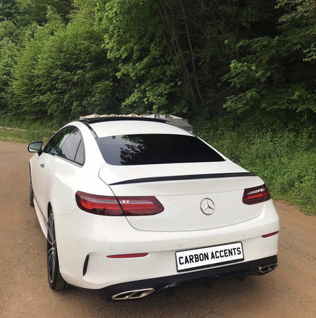 A white Mercedes-Benz coupe is parked on a dirt road, surrounded by lush green trees. The license plate reads "CARBON ACCENTS."