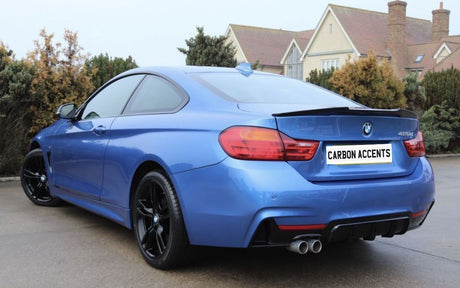 Blue sports car parked on a pavement, displaying dual exhausts and black rims. Background features a row of shrubs and residential houses. License plate reads "CARBON ACCENTS."