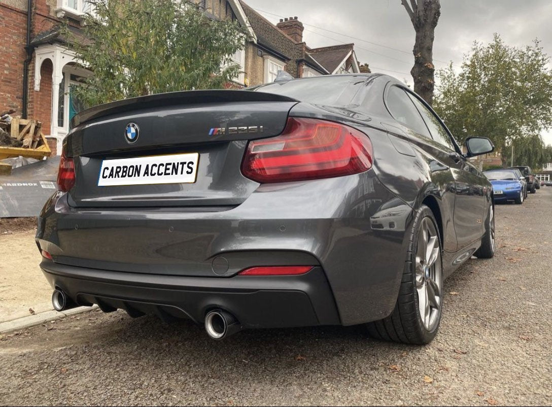 A gray BMW M235i parked on a residential street, showcasing dual exhausts and a rear spoiler. The license plate reads "CARBON ACCENTS." Nearby, brick houses and trees line the street.