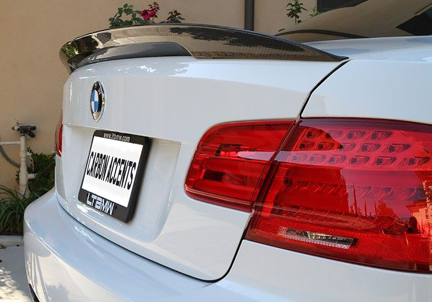 A white BMW features a dark carbon fiber spoiler. The rear view reveals red tail lights and a license plate frame displaying "CARBON ACCENTS" with "LTBMW" and "www.ltmbmw.com" below.