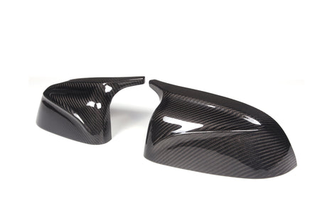 Two carbon fiber mirror covers, glossy with a black and gray weave pattern, are positioned against a plain white background. They exhibit a sleek, aerodynamic design.