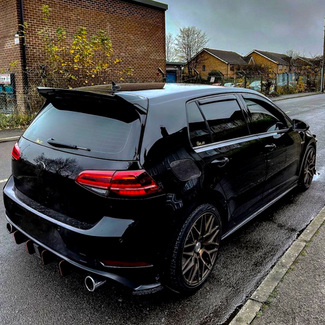 A black hatchback car is parked on a wet street, featuring tinted windows and dual exhaust pipes. It is adjacent to a brick building and some bushes under an overcast sky.