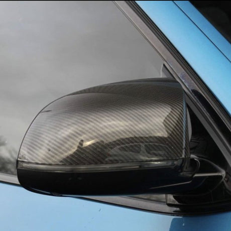 A carbon fiber car side mirror reflects light, attached to a blue vehicle door, against a backdrop of a cloudy sky and faint reflections of surrounding trees.