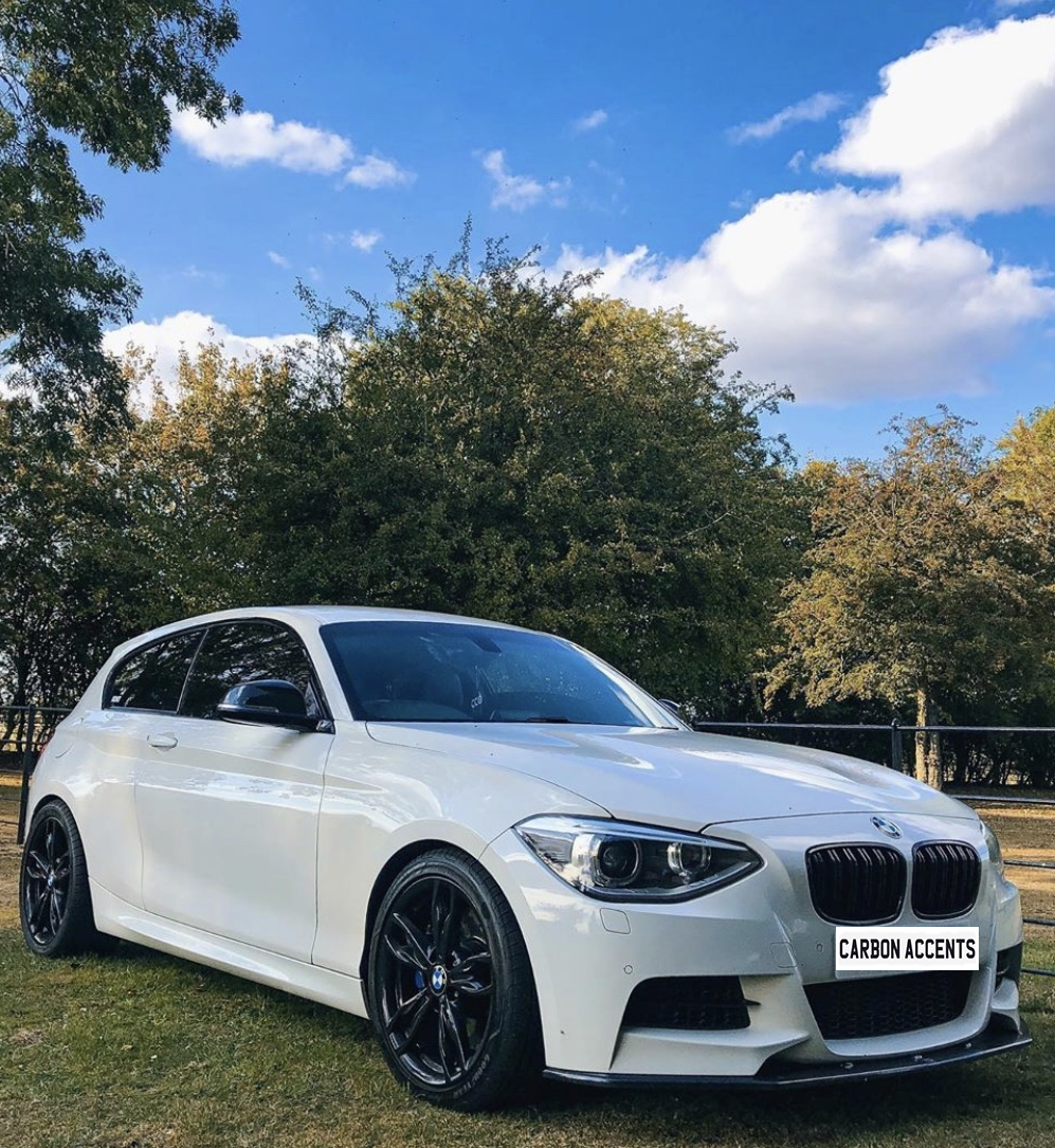 A white car with black rims is parked on grass, surrounded by trees under a blue sky with clouds. Text on the license plate reads "CARBON ACCENTS."