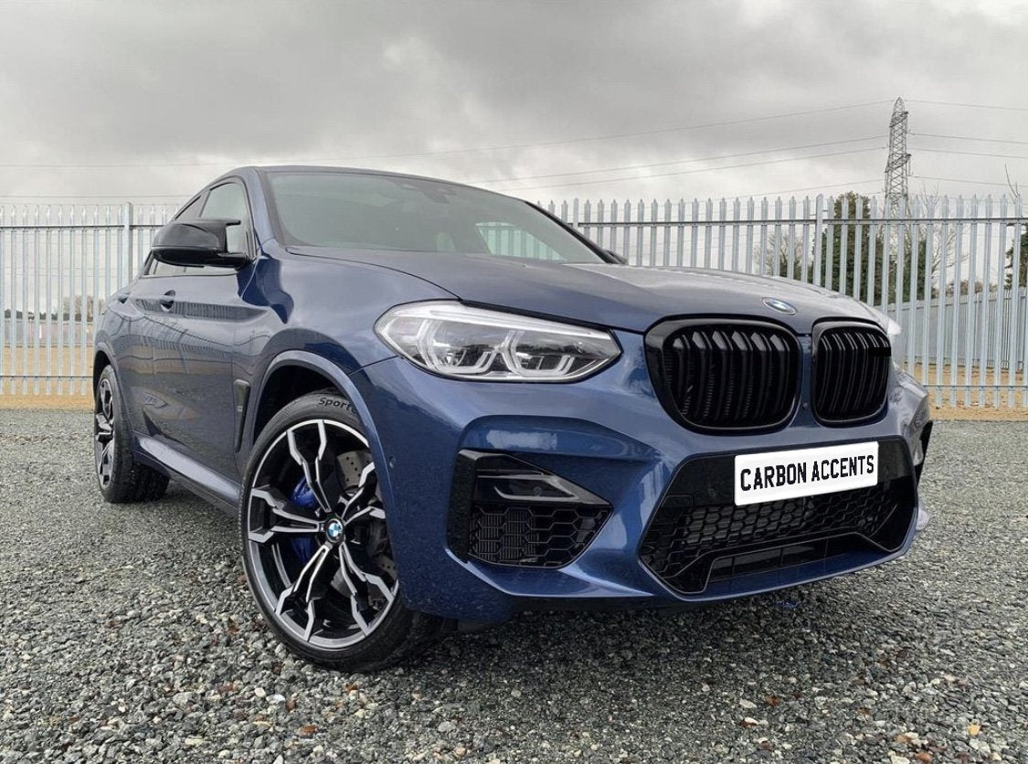 A blue SUV with a sleek design is parked on gravel, featuring stylish alloy wheels and the text "CARBON ACCENTS" on the front plate, set against a cloudy sky and metal fencing.