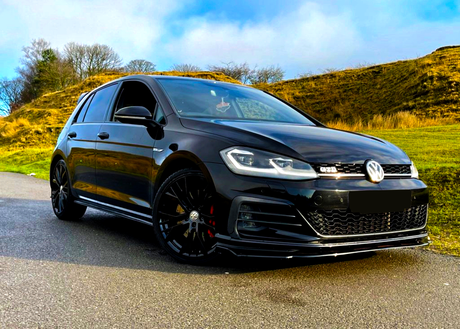 A black car is parked on a road, facing slightly left. It features "GTD" on the front grill, surrounded by grassy hills and bare trees under a blue sky.