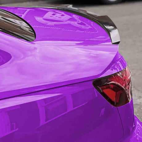 A vibrant purple car features a sleek, tinted taillight and a glossy black spoiler. It is parked on an asphalt street, reflecting nearby buildings.