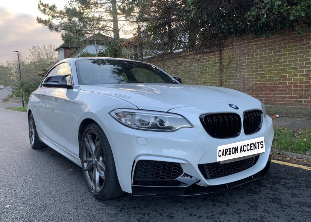 A white BMW parked on a quiet street, with trees and a brick wall in the background. The license plate reads "CARBON ACCENTS."