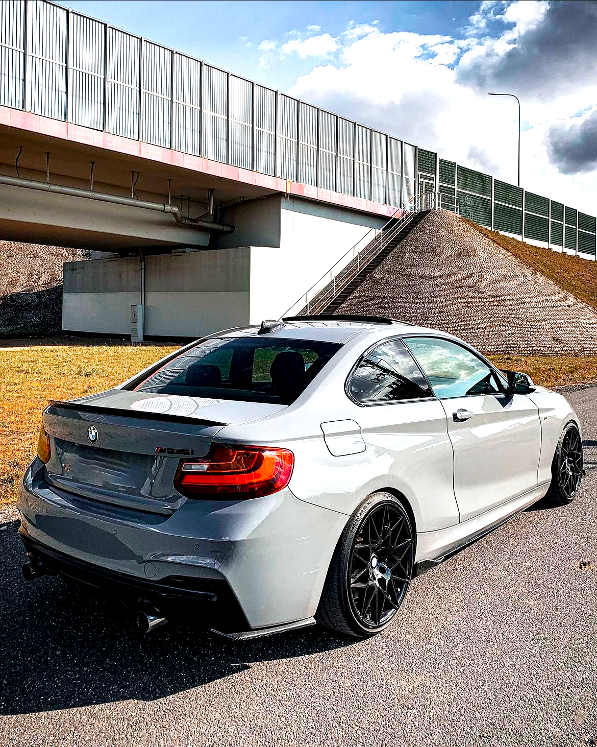 A gray BMW M235i is parked on a paved road beside a grassy mound and under a bridge, with a cloudy sky above.