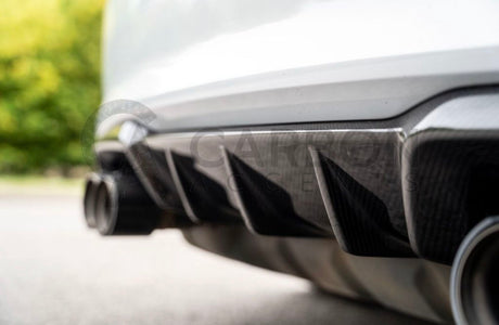 Car rear diffuser with exhaust pipes, surrounded by blurred greenery and pavement. Text reads: "CARBON ACCENTS," faintly visible over the image in gray.