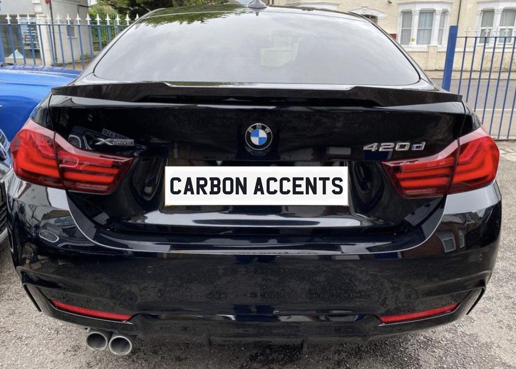 The black BMW 420d is parked, displaying "CARBON ACCENTS" on its license plate. It’s positioned in an outdoor setting with a blue car and a building nearby.