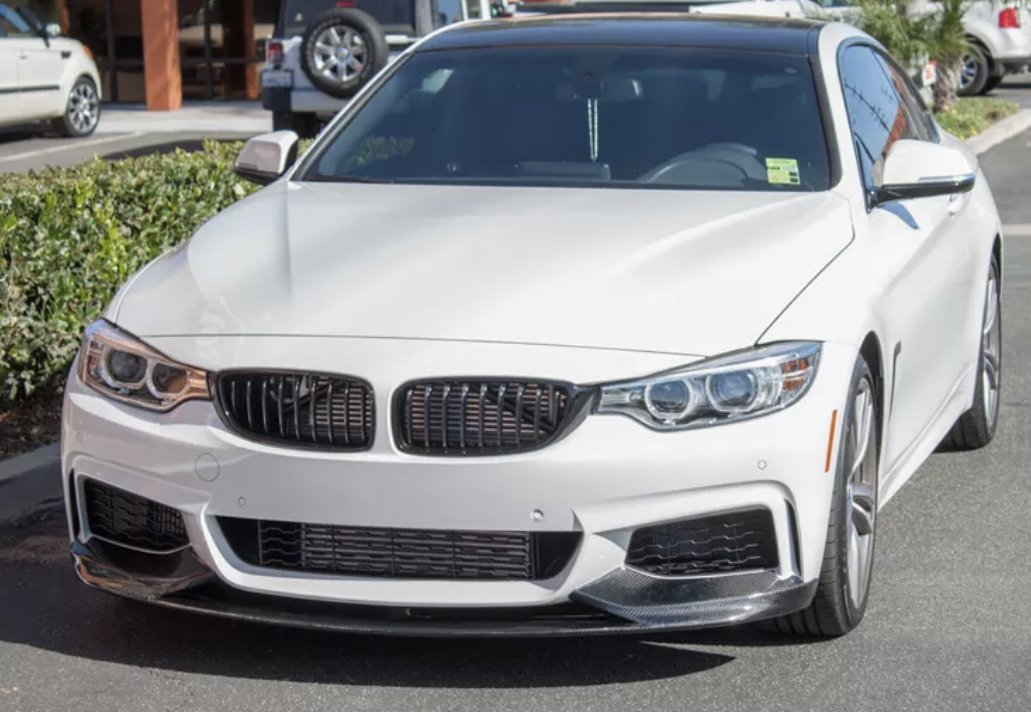 White car parked in a lot, with a sleek design featuring a prominent grille and headlights. Bushes and another vehicle are visible in the background under clear daylight.