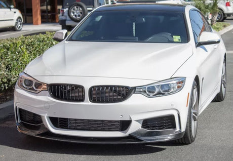 White car parked in a lot, with a sleek design featuring a prominent grille and headlights. Bushes and another vehicle are visible in the background under clear daylight.