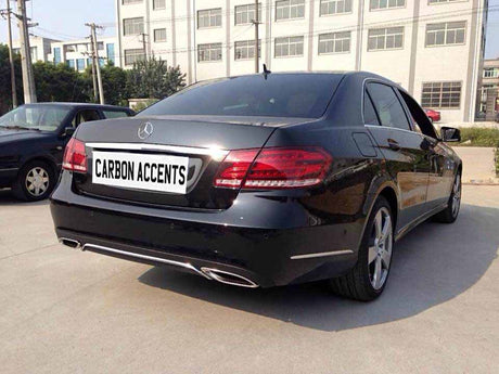 Car stationary in a parking lot, displaying a "CARBON ACCENTS" license plate. Nearby, another car is parked. Background features a multi-story industrial building and trees.