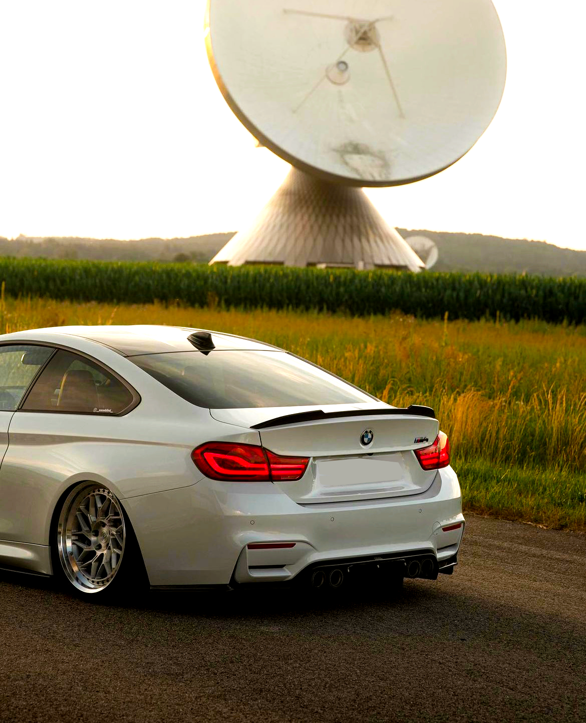 A white car with glossy rims is parked on a road near tall grass, while a large satellite dish looms in the background against a sunset sky.