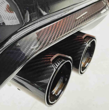 Dual exhaust pipes with a sleek carbon fiber finish emerge from a vehicle's rear bumper. The setting is a clean, bright environment, enhancing the contrast and details.