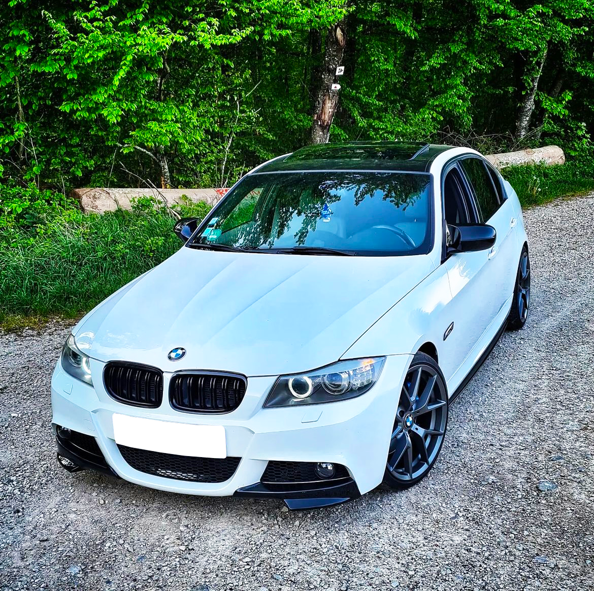 A white BMW car is parked on a gravel path against a backdrop of lush greenery, with sunlight filtering through the trees.
