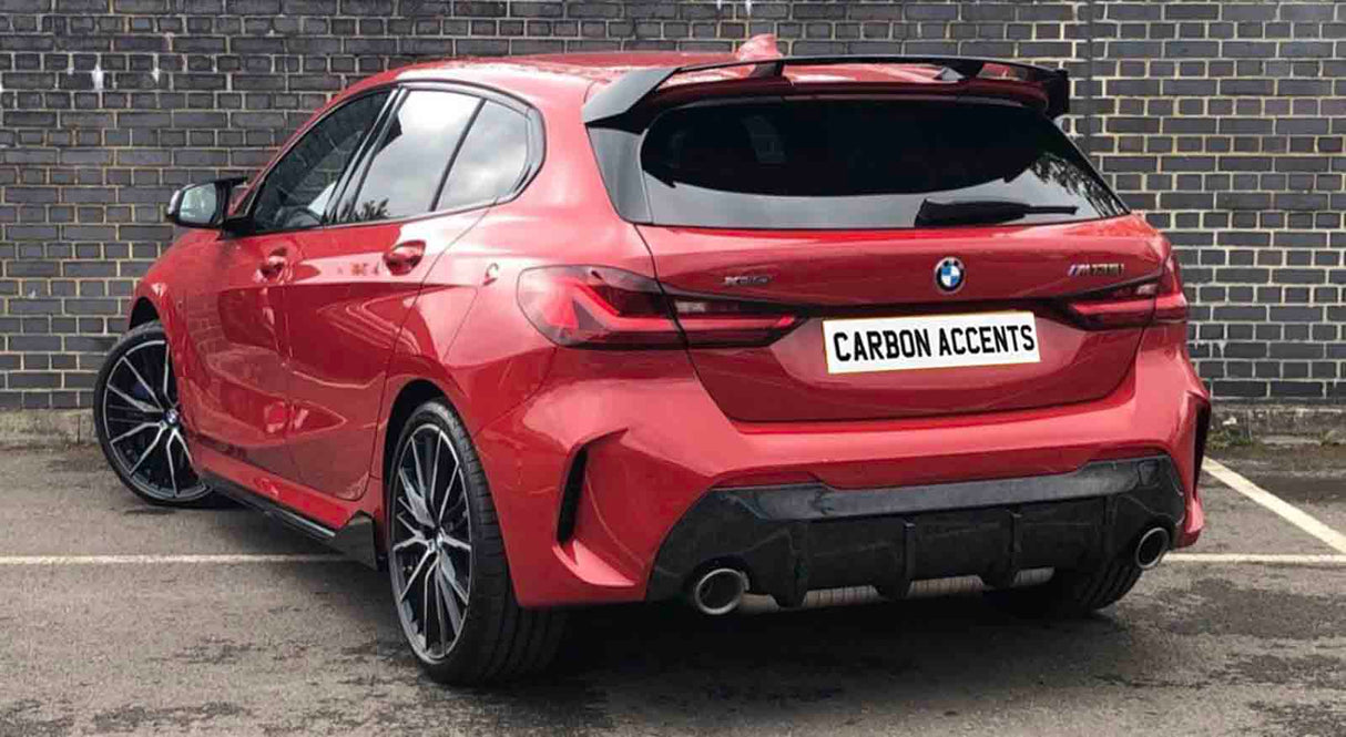 A red BMW hatchback, parked by a brick wall, displays "CARBON ACCENTS" on the license plate. The vehicle is stationary, showcasing sleek, modern design and large wheels.