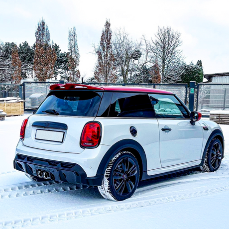 A white car with a red roof is parked on snow, leaving tire tracks. It's in a fenced area with trees and a building in the background.
