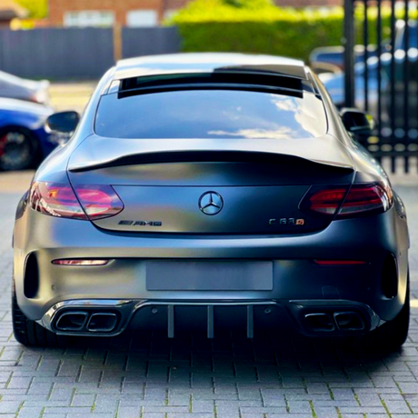 A matte black Mercedes-Benz AMG C63 parked on a paved surface, features visible exhausts and logos. Background includes parked cars and blurred greenery with a fence.
