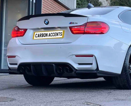 A white BMW car, parked outdoors, displays a rear spoiler and quad exhausts. The license plate reads "CARBON ACCENTS." The background includes a building entrance and greenery.