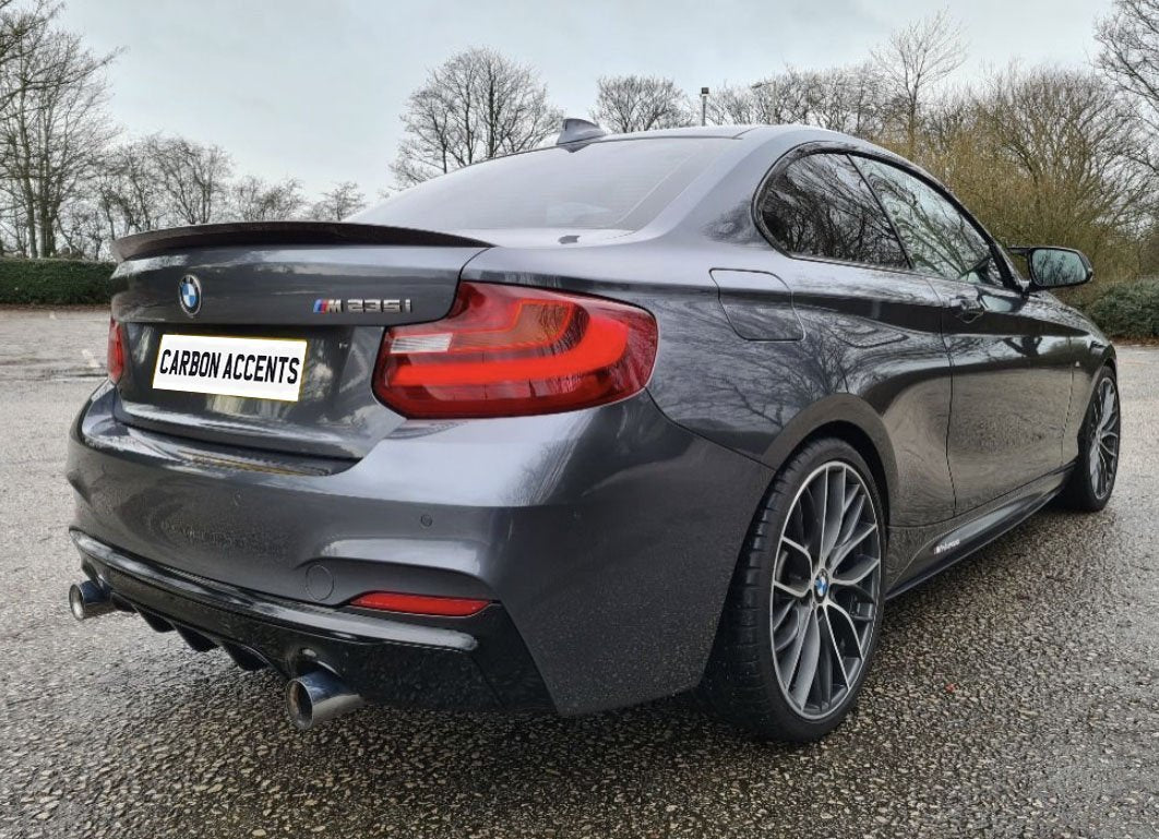 A dark gray BMW M235i is parked on a wet, textured ground with barren trees in the background. The license plate reads "CARBON ACCENTS."