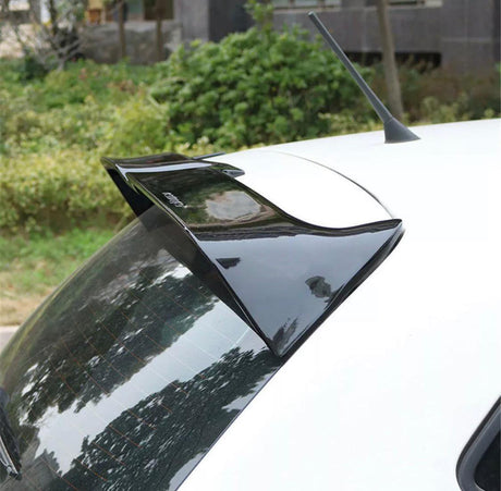 A black car spoiler is attached to the roof of a white vehicle, parked outdoors near green bushes and trees, with the word "Cuerpo" printed on the spoiler.