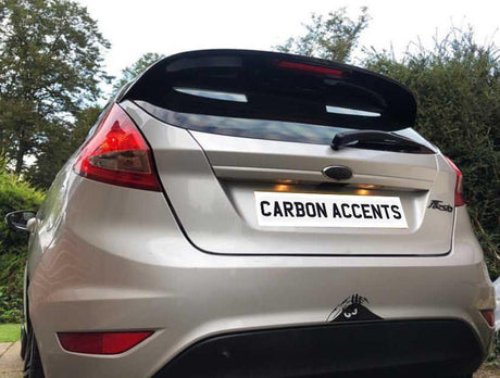 A silver car with a rear spoiler and "CARBON ACCENTS" on the rear plate is parked in a driveway surrounded by trees and bushes.