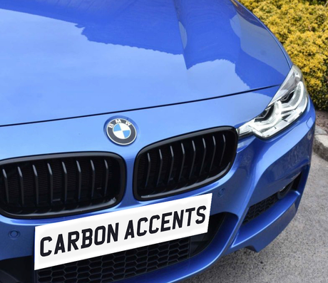 A blue BMW car with a prominent front grille and headlights is parked on a paved surface next to bushes. Text on the grille reads "CARBON ACCENTS."
