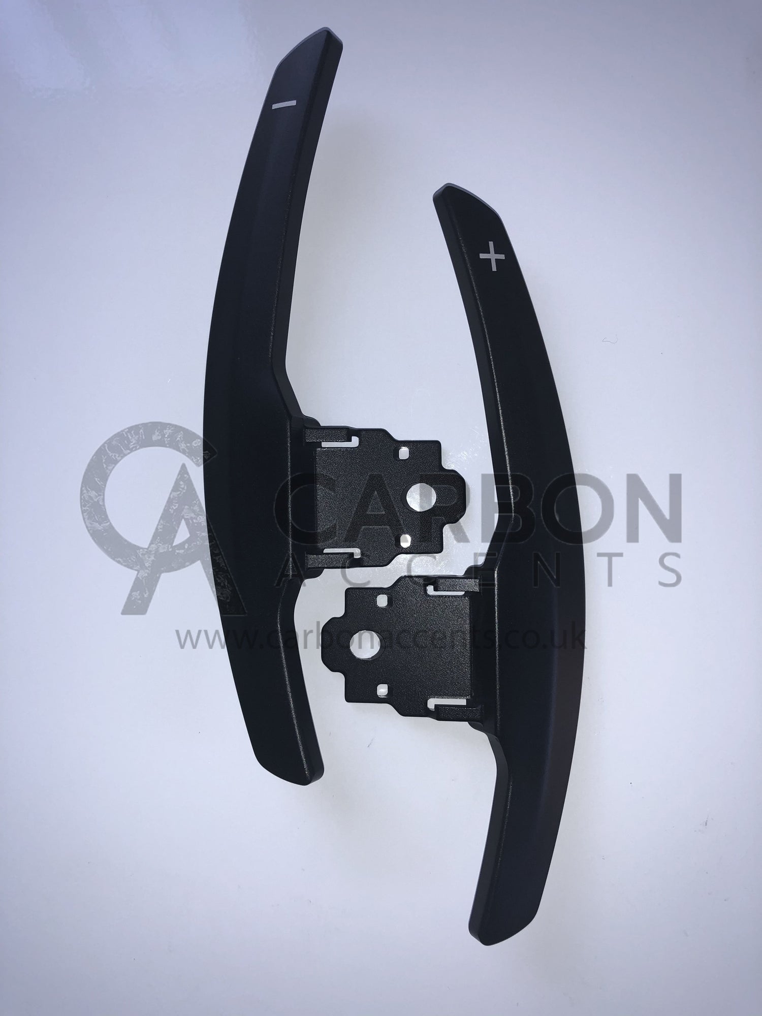 Two black paddle shifters with "+" and "-" symbols lie on a white surface. The background features text: "CARBON ACCENTS www.carbonaccents.co.uk."