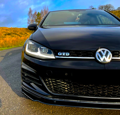 A black Volkswagen car with a "GTD" badge is parked on an asphalt road, surrounded by golden grass and trees under a blue sky.