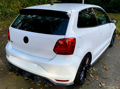 A white hatchback car with dark tinted windows is parked on a leaf-strewn asphalt path, surrounded by trees. The car features a rear spoiler and red taillights.
