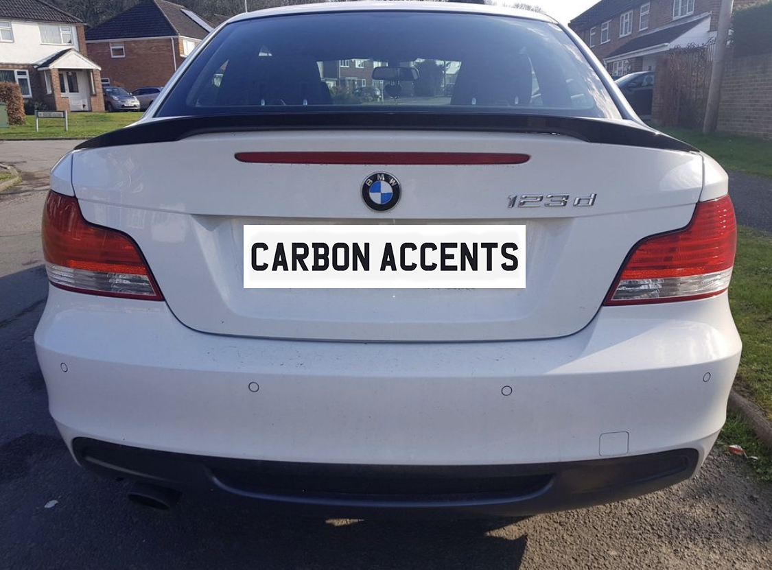 A white BMW 123d is parked in a residential neighborhood. The rear license plate reads "CARBON ACCENTS," with neighboring houses visible in the background.