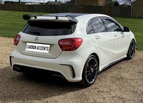 White hatchback car parked on gravel, with black rims and spoiler, viewed from the rear in a grassy area. License plate reads "CARBON ACCENTS."