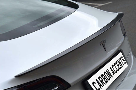 Car trunk with carbon fiber spoiler parked on asphalt, displaying logo and text "CARBON ACCENTS" on license plate. The rear design is sleek and modern.