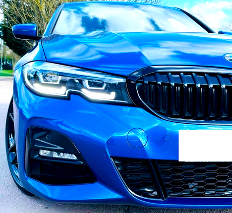 A blue car with illuminated headlights and a black grille is parked on a road, with trees and a cloudy sky visible in the background.