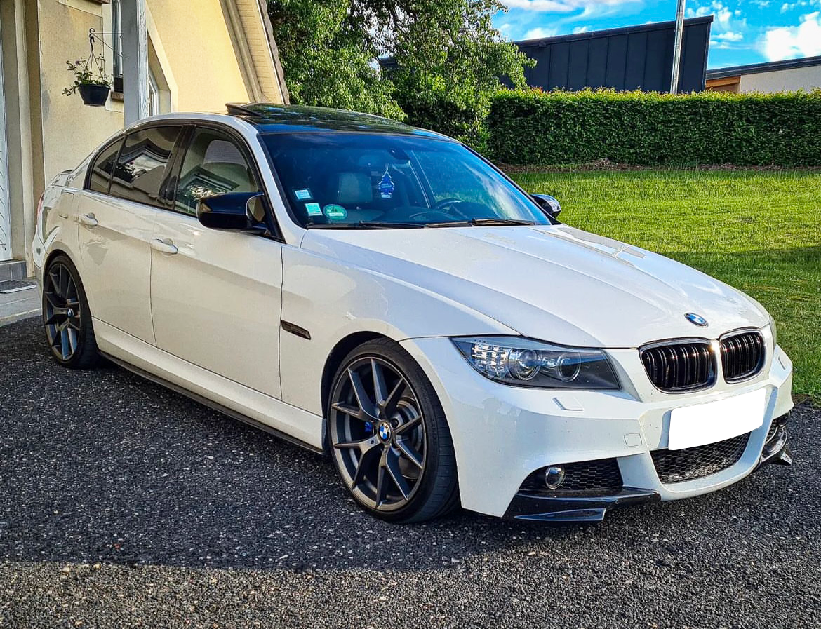 A white BMW sedan is parked on a paved driveway, next to a house with green grass and trees in the background under a partly cloudy sky.