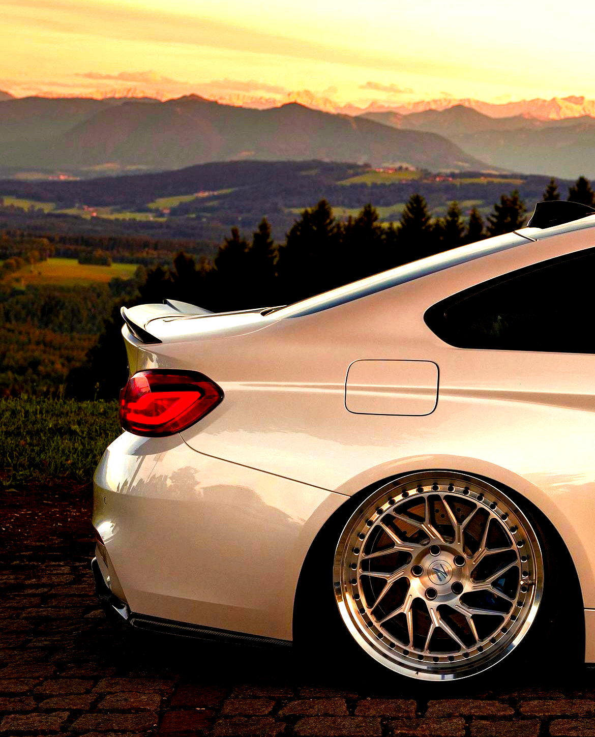 A sleek, silver sports car is parked on cobblestone, showcasing its shiny alloy wheels. It is set against a scenic backdrop of rolling hills and a sunset-lit mountain range.