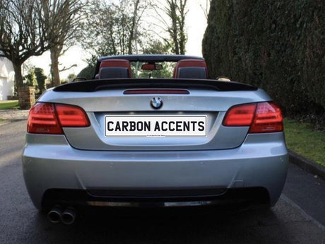 Convertible car parked on a residential street, viewed from the rear. The license plate reads "CARBON ACCENTS." Trees and hedges line the background.
