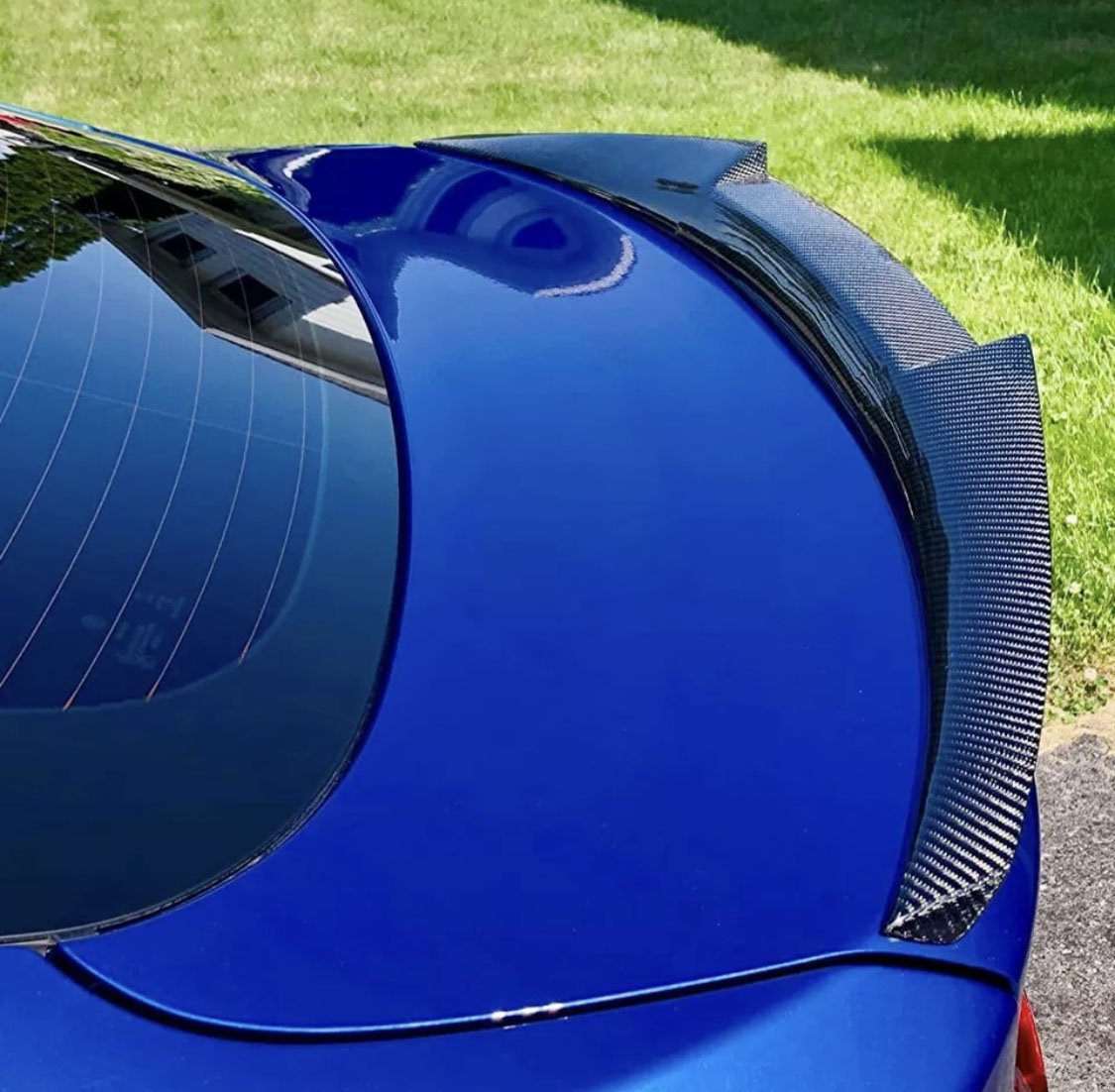 A blue car with a carbon fiber spoiler reflects sunlight, parked on grass. The rear window shows building reflections, emphasizing the car's glossy finish and sporty design.