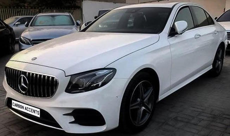 A white Mercedes-Benz sedan is parked on a concrete pavement. Its front grille displays the Mercedes emblem. The vehicle is surrounded by other parked cars. The license plate reads "CARBON ACCENTS."