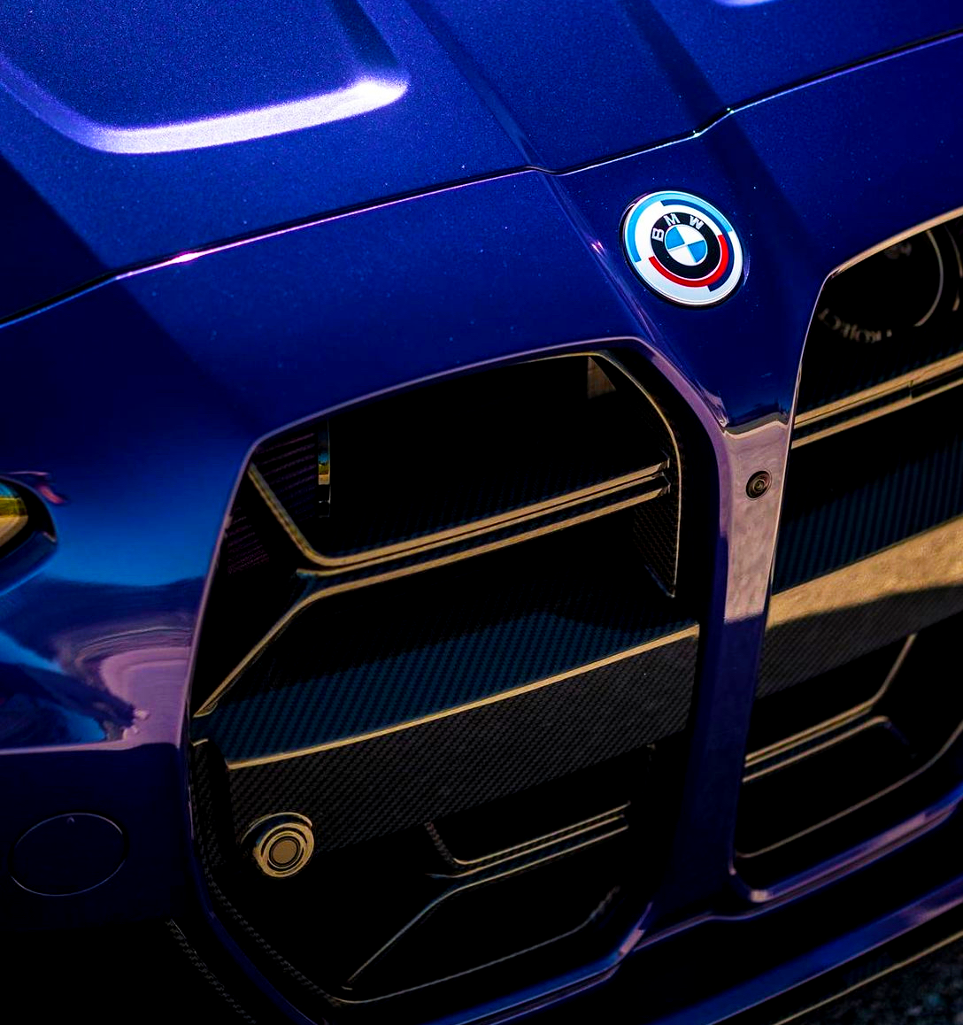 A blue car front displays a prominent grille and a round BMW emblem above it, situated outdoors in bright sunlight.