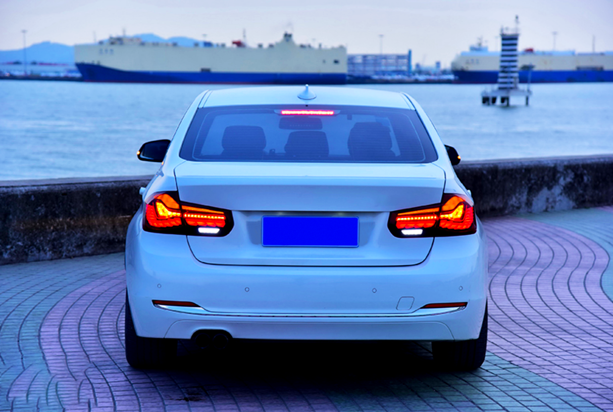 A white car is parked with illuminated taillights, facing a calm waterfront. In the background, large ships and a distant horizon are visible under a clear sky.