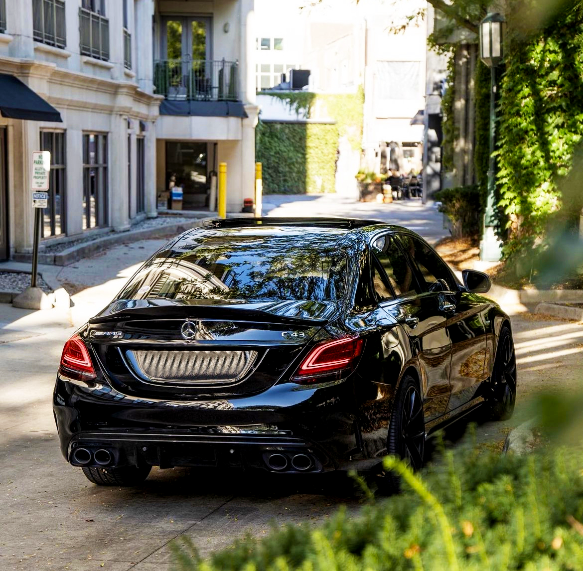 A black Mercedes-Benz C43 car is parked on a narrow, sunlit street lined with buildings and greenery. A sign reads "2 HOUR PARKING CUSTOMER PARKING ONLY."